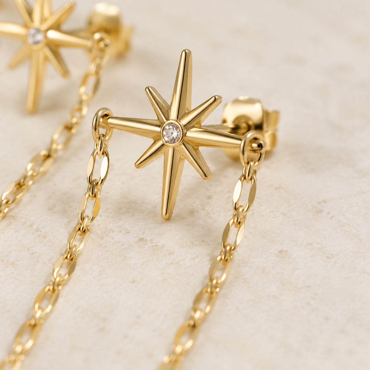 Gold starburst stud earrings with a small clear gemstone in the center, attached to delicate gold chains, displayed on a light beige textured surface.