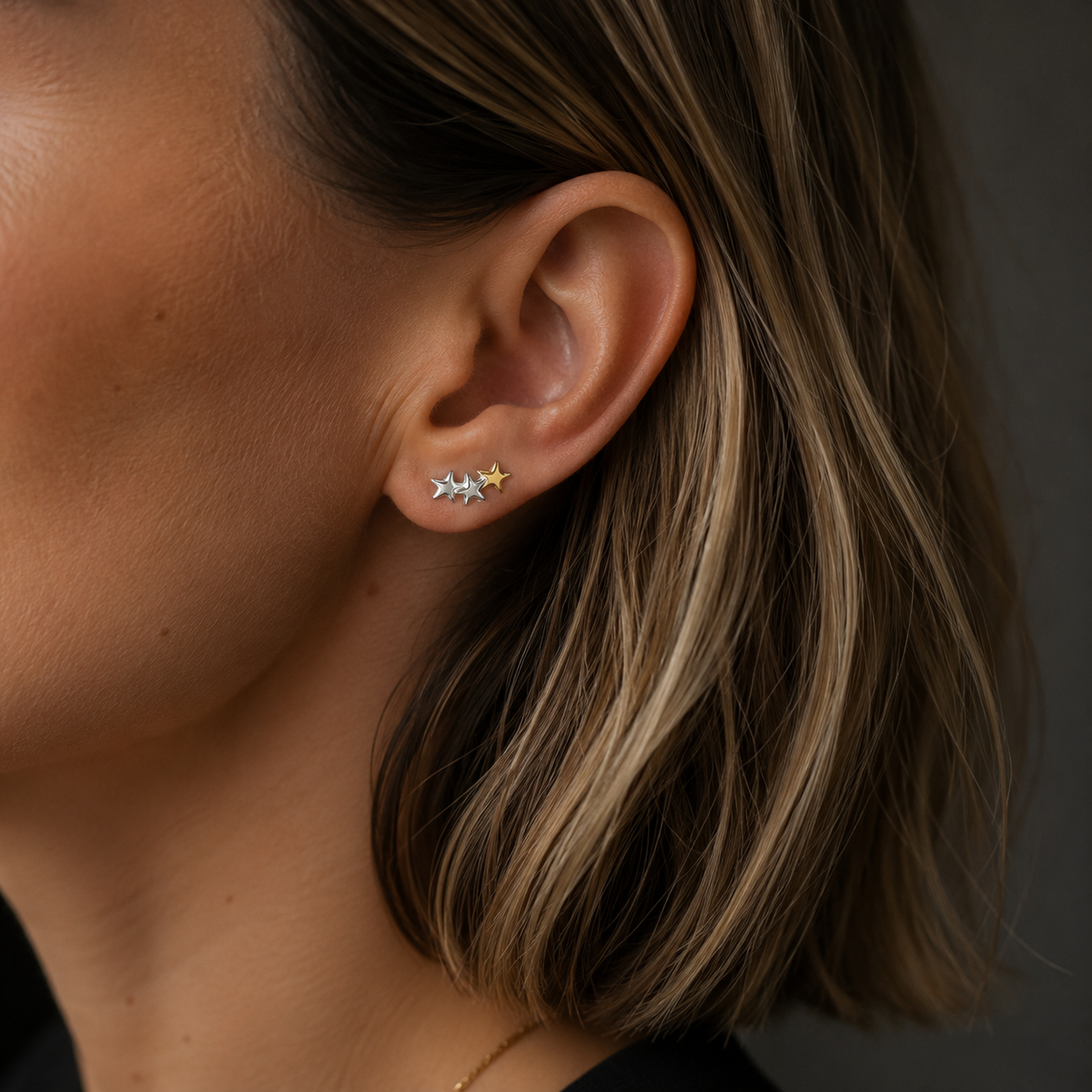 A close-up of a woman's ear wearing a small cluster stud earring with three tiny star-shaped settings, one in yellow gold and two in silver.