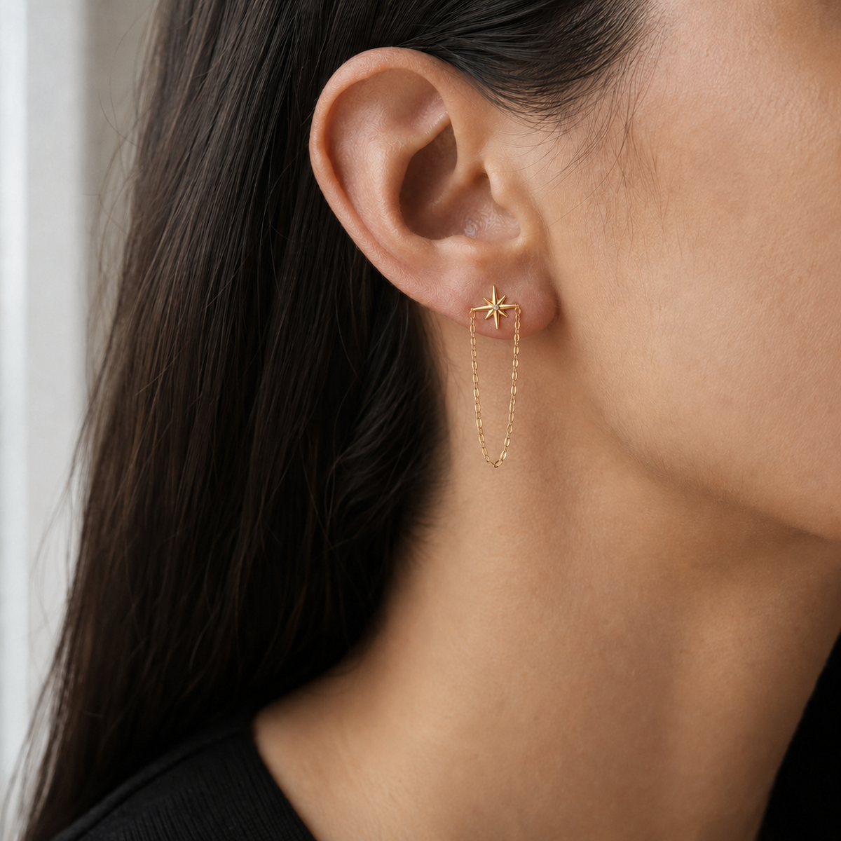 A close-up of a model's ear wearing a delicate gold starburst stud earring with a long dangling chain.
