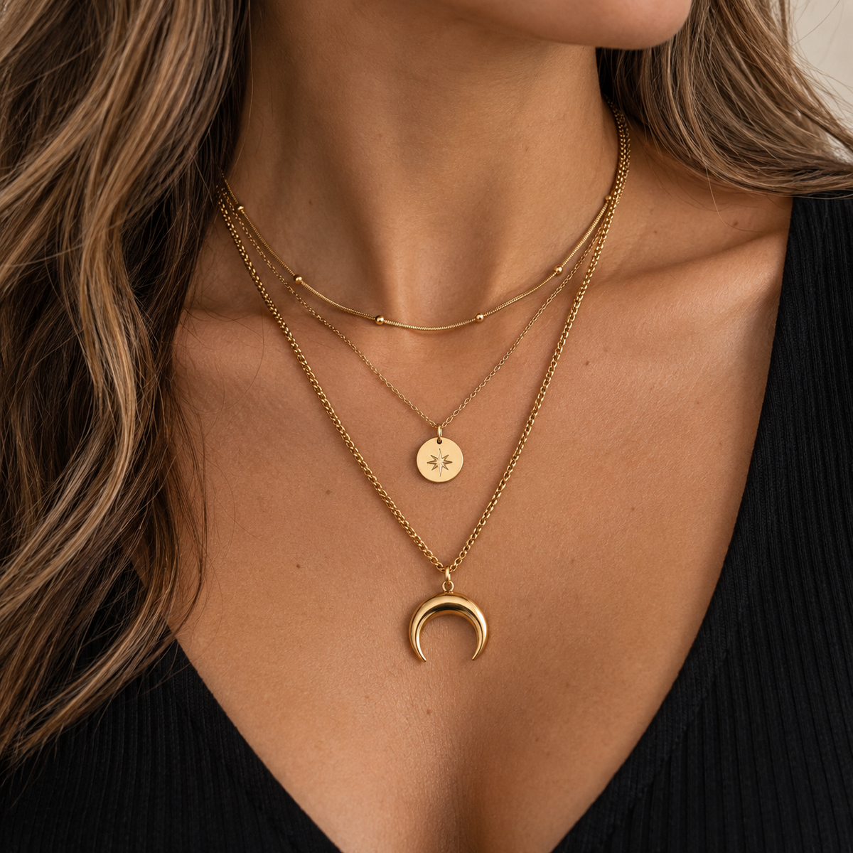 A close-up of a model's neck and chest wearing three layered gold necklaces: a short curb chain, a longer chain with a small round star disc pendant, and a longer chain with a crescent moon pendant.