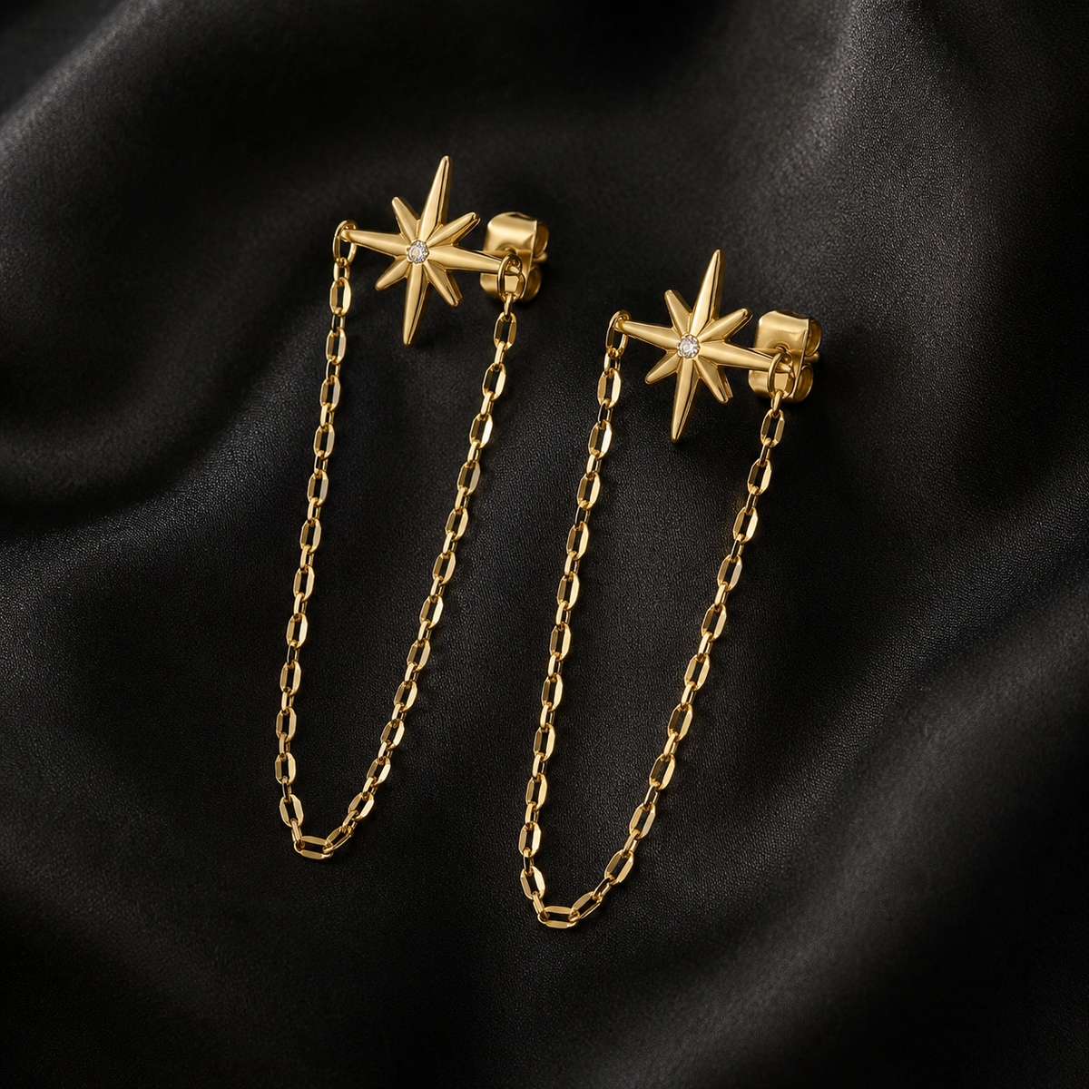 A pair of gold-toned starburst stud earrings with delicate chain drops, displayed on a draped black satin fabric background.
