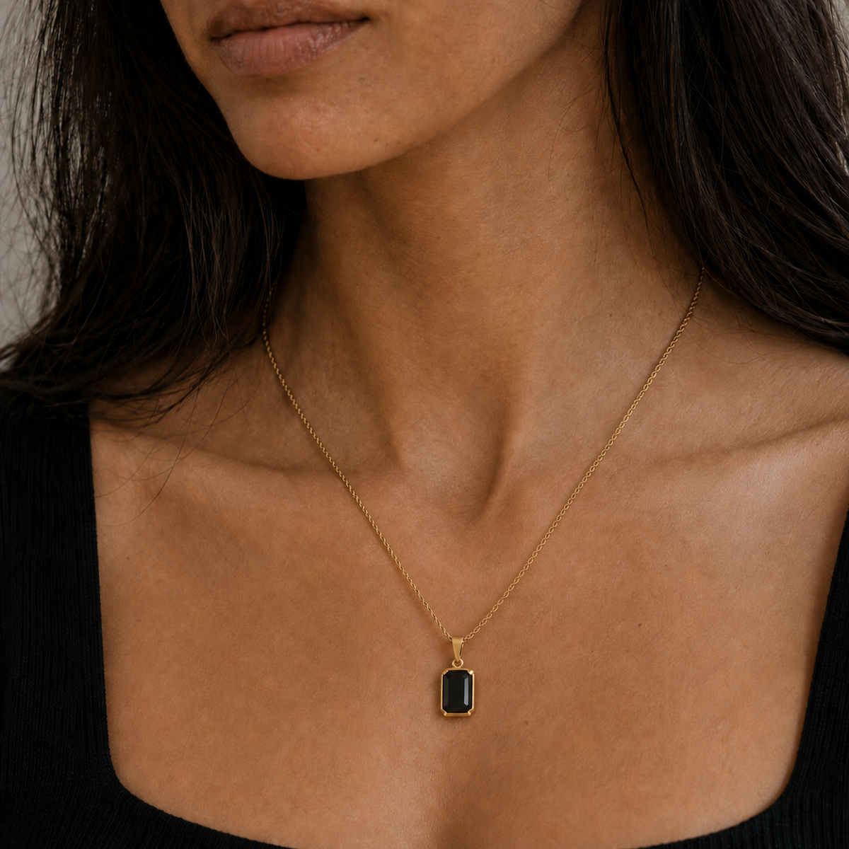 A close-up of a woman wearing a delicate gold chain necklace with a small rectangular black pendant, shown against her neck and upper chest.