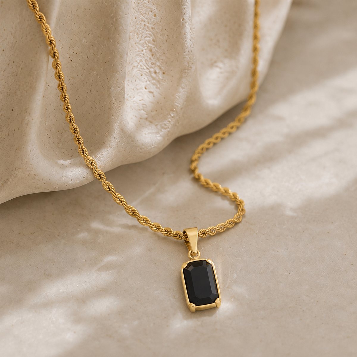 A gold rope chain necklace with a rectangular black gemstone pendant, styled on a textured beige surface with draped cream fabric.