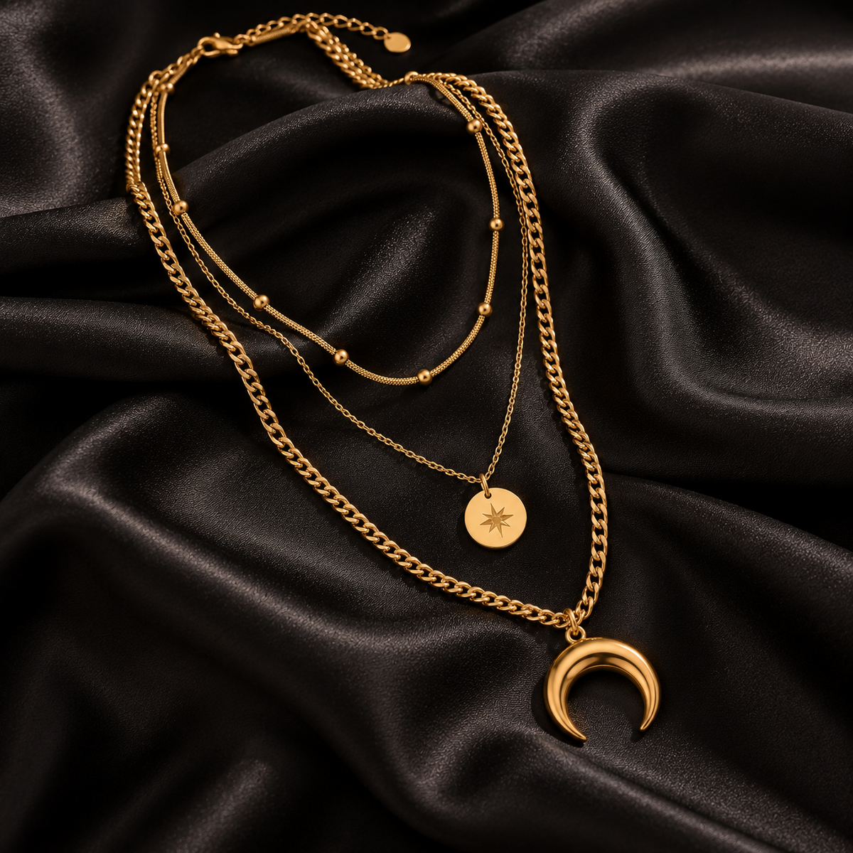 A layered gold necklace set displayed on draped black satin fabric. The set includes a long curb chain, a beaded chain, a short chain with a round starburst medallion, and a larger crescent moon pendant.