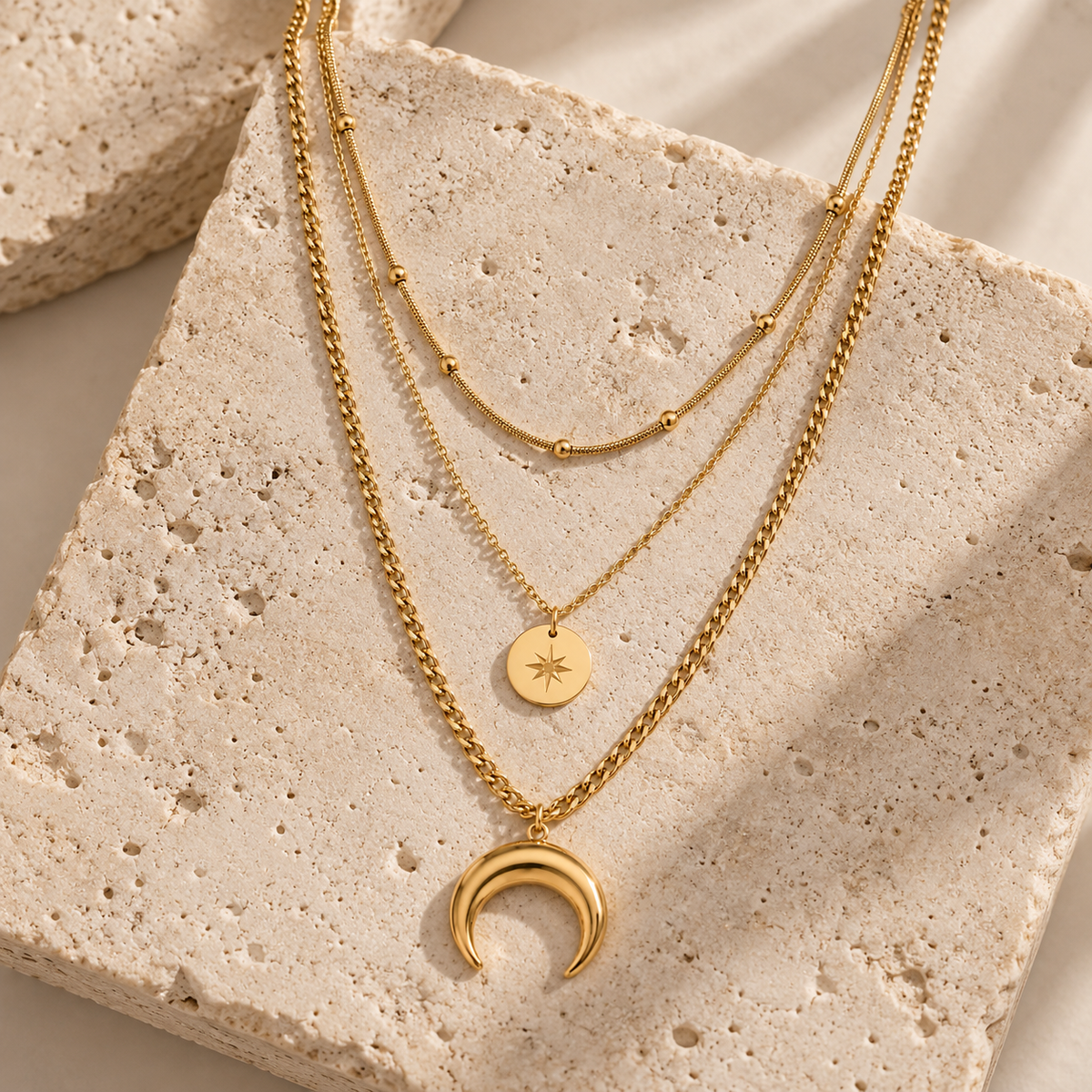Three layered gold necklaces displayed on a textured beige stone slab. The necklaces feature different chain styles and pendants: a delicate chain with small beads, a chain with a round star disc pendant, and a chain with a crescent moon pendant.