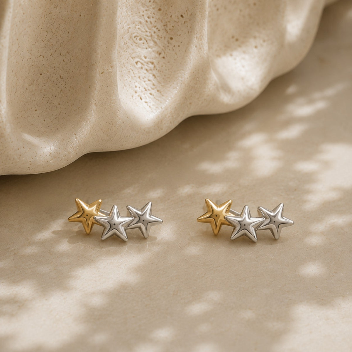 A pair of small star-shaped stud earrings in mixed gold and silver tones, displayed on a warm beige surface with a sculptural beige prop and soft dappled shadows.